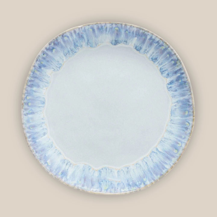 A round ceramic plate with a smooth, pale blue center and a wide, textured outer rim featuring blue, white, and light gray marbled patterns, shown against a solid beige background.