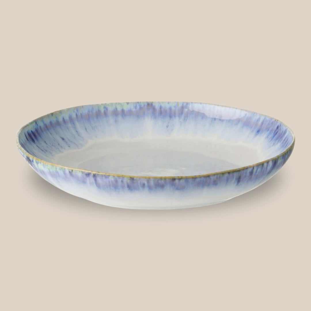 A shallow brisa serving bowl with a white interior and a blue reactive glazed exterior featuring a subtle rim, set against a solid beige background.