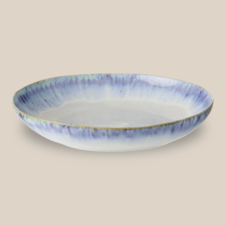 A shallow brisa serving bowl with a white interior and a blue reactive glazed exterior featuring a subtle rim, set against a solid beige background.