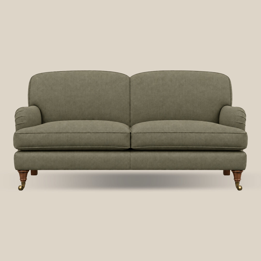 An upholstered Howard style sofa with two seat cushions and rounded arms, shown from the front against a plain warm beige background. The fabric is a muted sage green, and the legs are dark wood with brass casters.