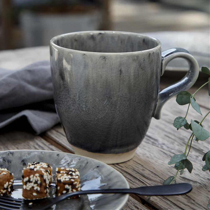 Grey ceramic mug on a wooden table with a plate of pastries and a plant.